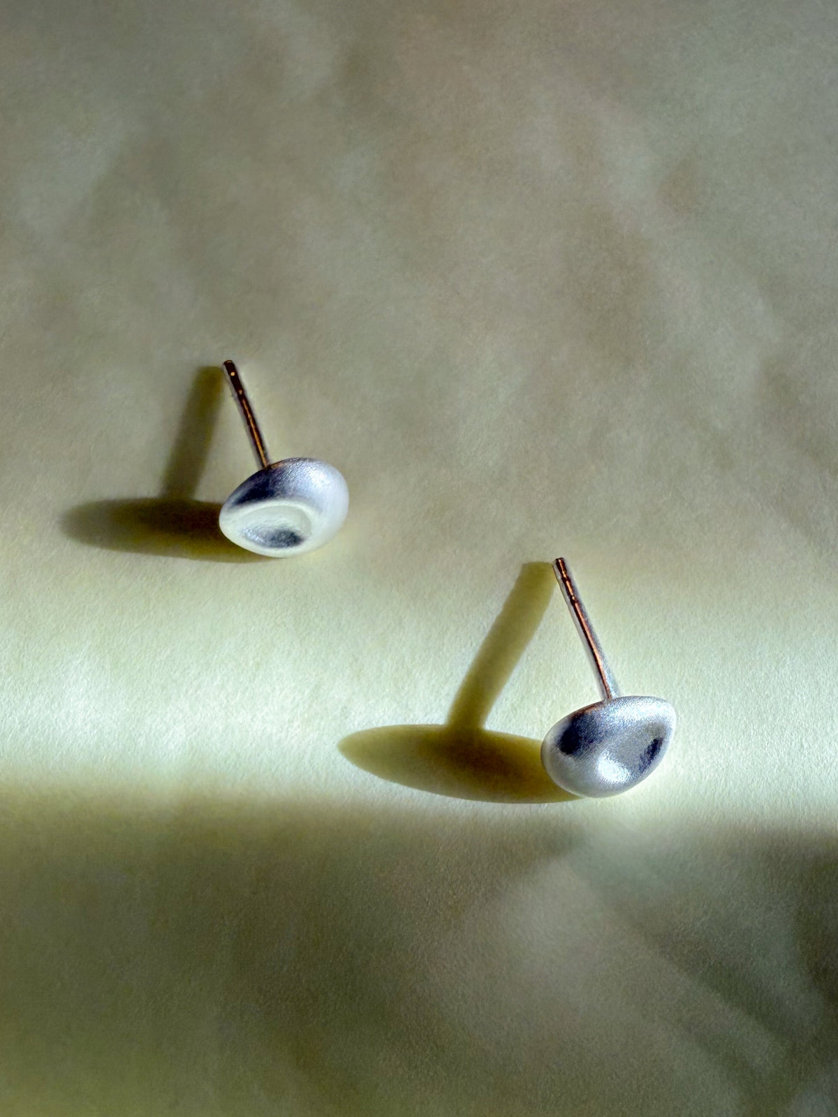 A pair of silver dome stud earrings with rose gold posts, resting on a textured beige surface with dramatic shadows.