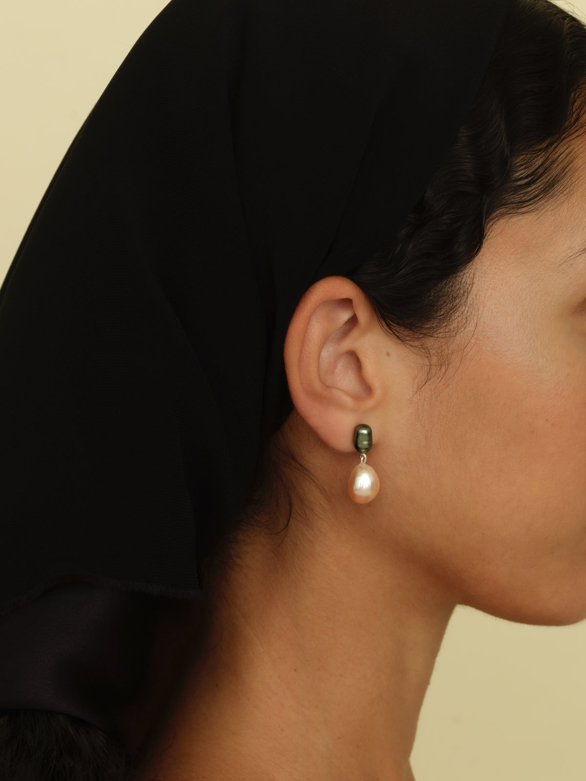 A close-up of a model's profile wearing a single drop earring with a small dark green bead and a teardrop-shaped soft pink pearl and a black silk scarf.