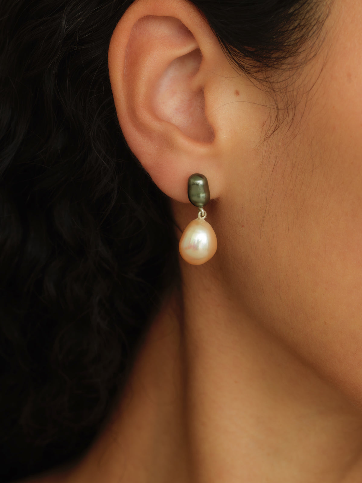 A close-up of a person's ear wearing a drop earring with a dark olive-green top bead and a teardrop-shaped cream pearl dangling below.
