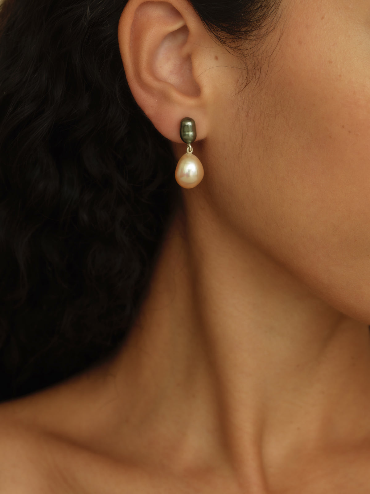 A close-up of a model's ear wearing a single drop earring with a small dark green bead and a teardrop-shaped soft pink pearl.