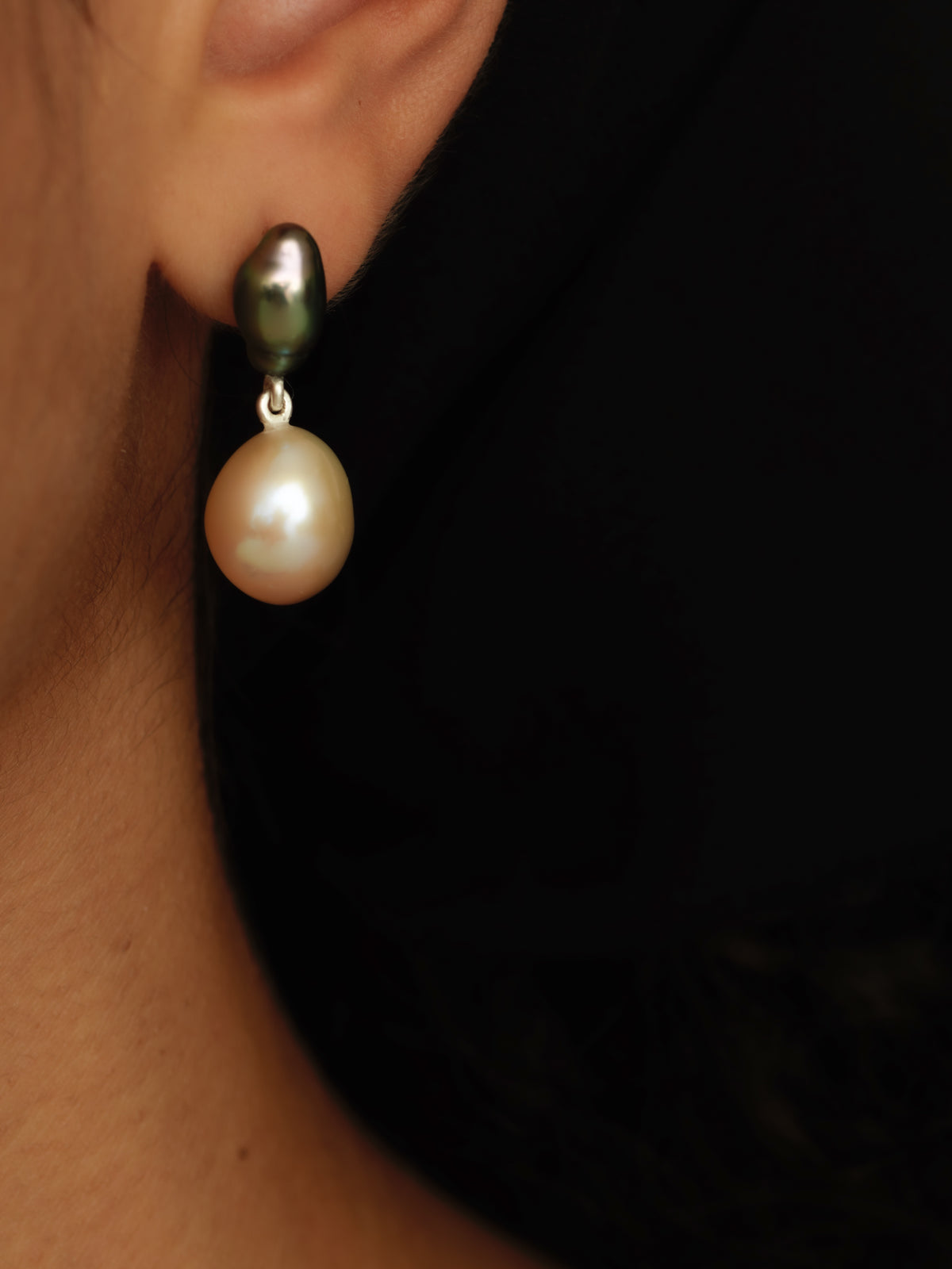 A close-up of a person's ear wearing a drop earring with a dark green-black baroque pearl and a larger soft pink freshwater pearl below in a silver setting.
