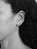 A close-up black and white photograph of a person's ear wearing a small round stud earring with a circular emblem.