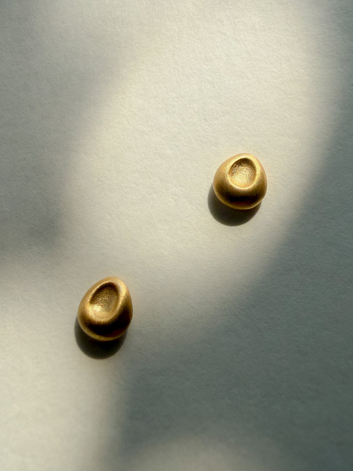 Two small gold oval stud earrings with a textured, dimpled surface, displayed on a light gray background with artistic shadows.