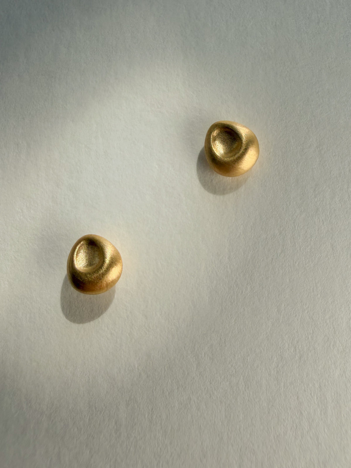 A pair of small, organic-shaped gold stud earrings with a textured, matte finish, displayed on a textured off-white paper background.