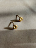 A pair of gold-toned stud earrings with a textured, organic circular face, resting on a beige linen fabric under warm, directional sunlight.