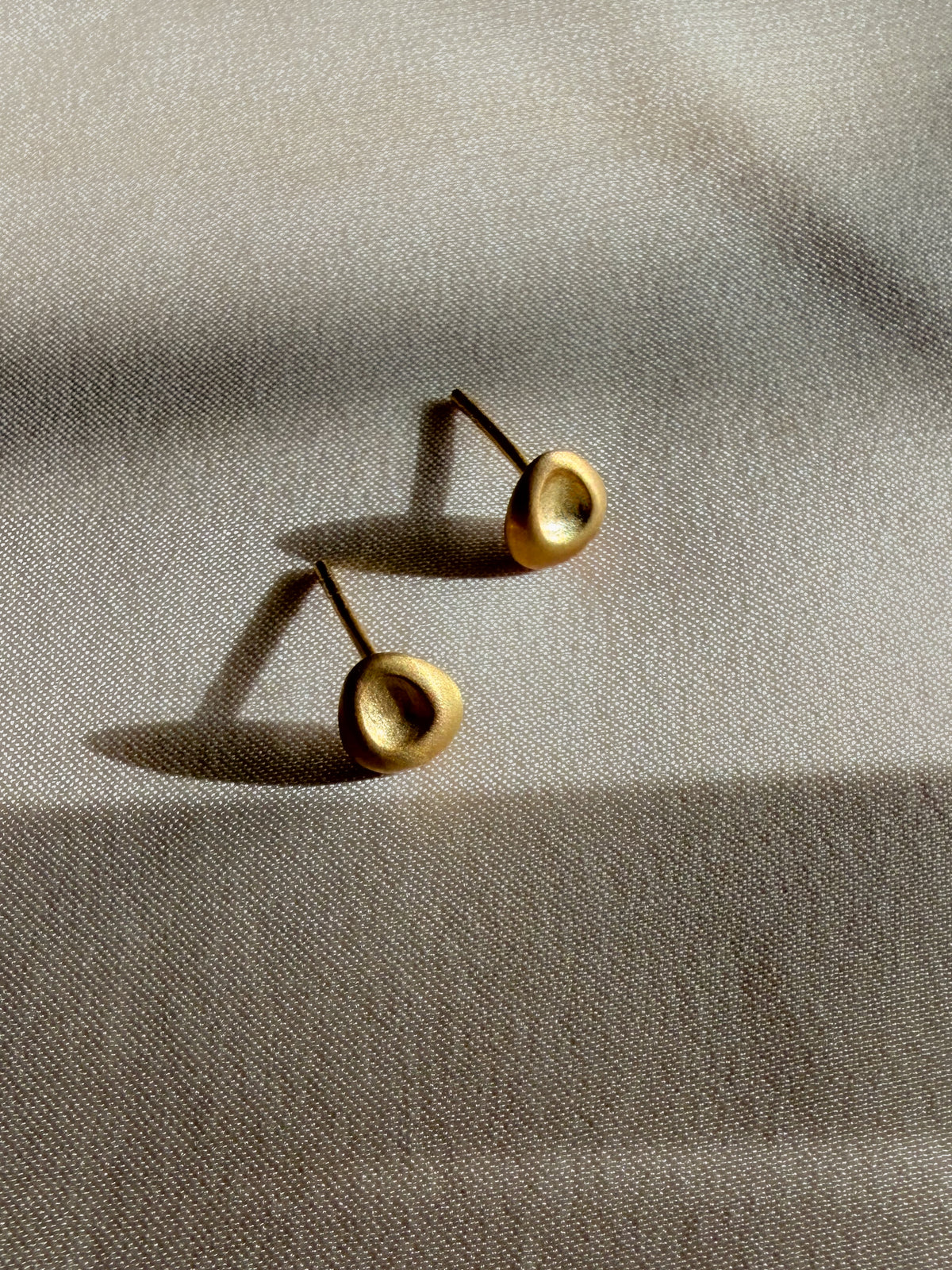 A pair of gold-toned stud earrings with a textured, organic circular face, resting on a beige linen fabric under warm, directional sunlight.