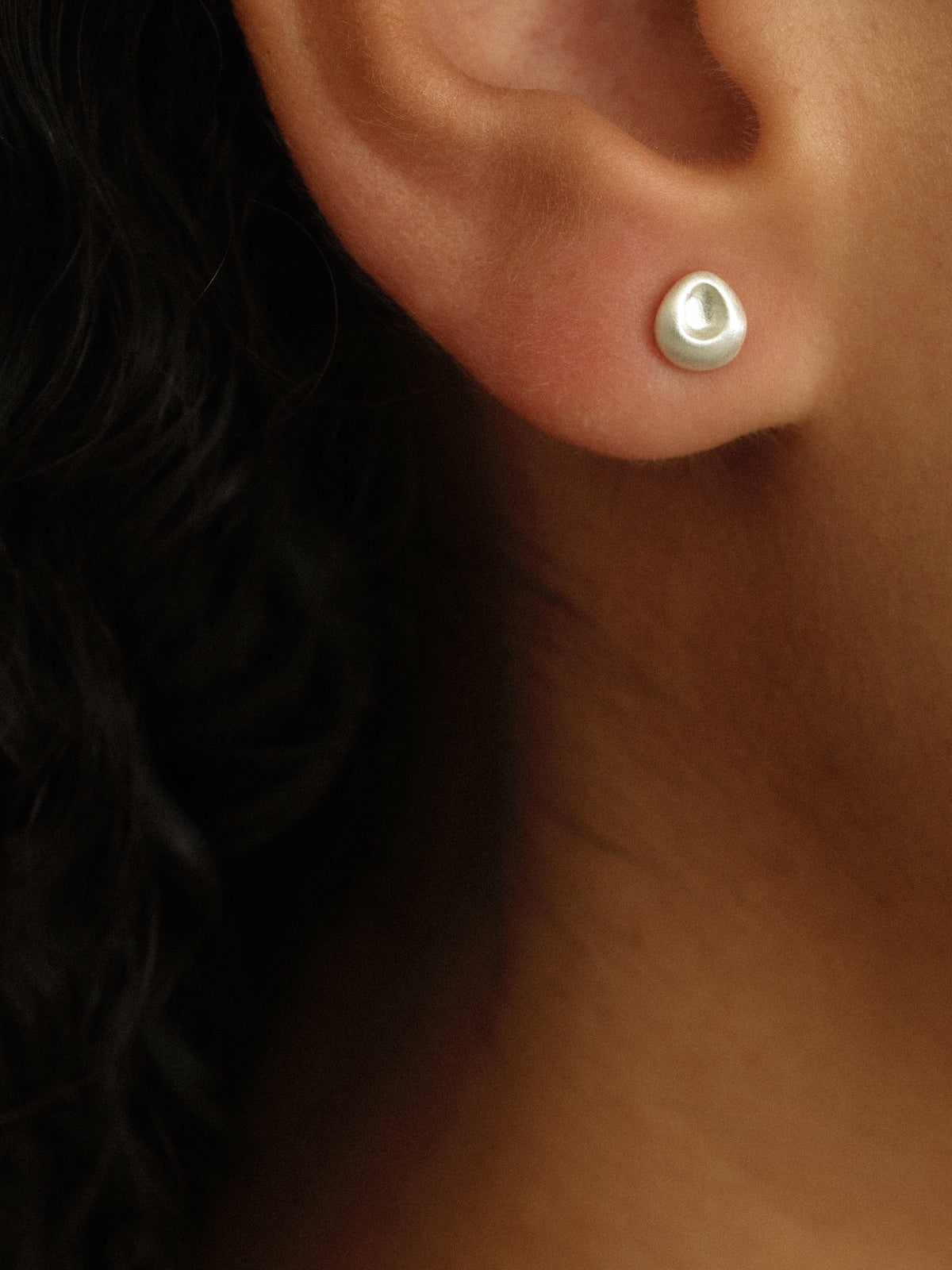 A close-up of a person's ear wearing a small, round, silver-toned stud earring with a polished, reflective surface.