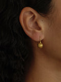 A close-up of a person's ear wearing a small gold teardrop dangle earring, with dark curly hair and skin visible.