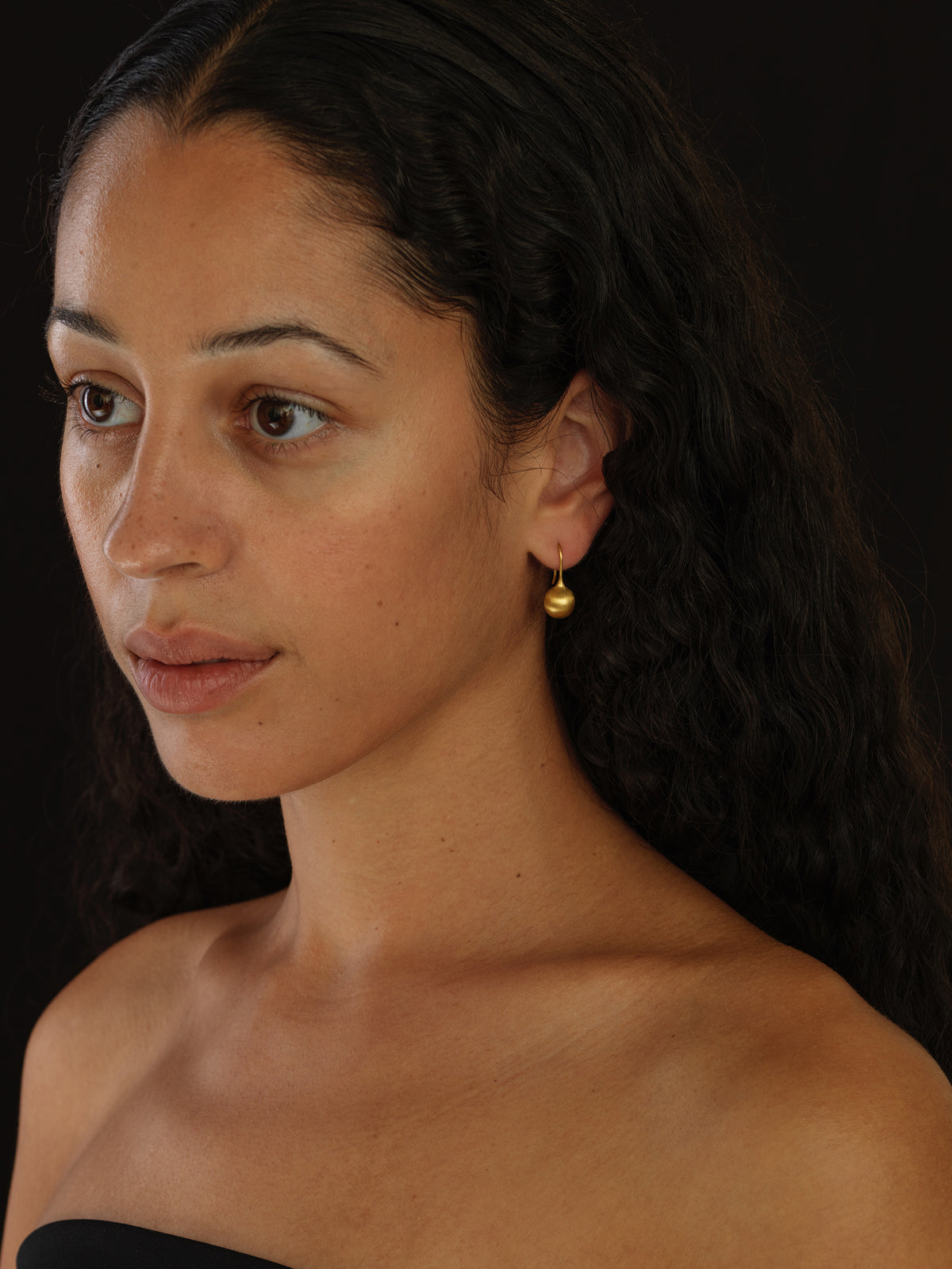 A close-up portrait of a woman with dark curly hair wearing a small gold hoop earring with a dangling gold bell, set against a dark background.
