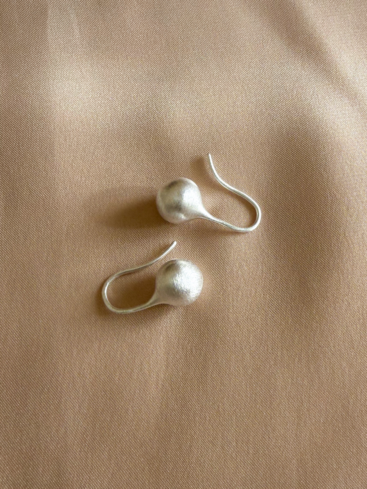 A pair of silver dangle earrings with textured spherical beads and fishhook backs, displayed on a beige fabric background.