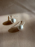 A pair of minimalist silver dangle earrings with textured white spherical beads, displayed on a beige fabric background with soft shadows.