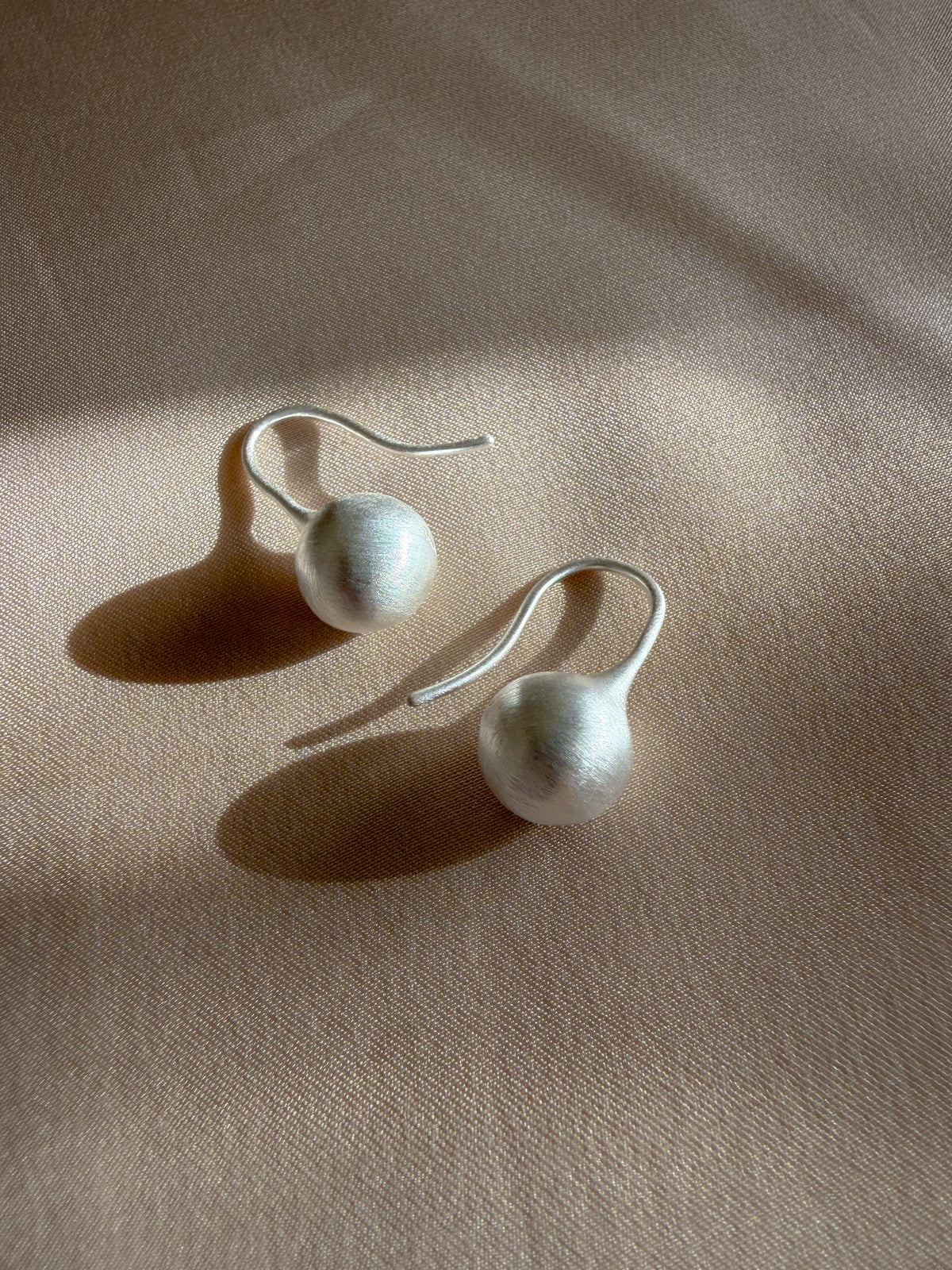 A pair of minimalist silver dangle earrings with textured white spherical beads, displayed on a beige fabric background with soft shadows.