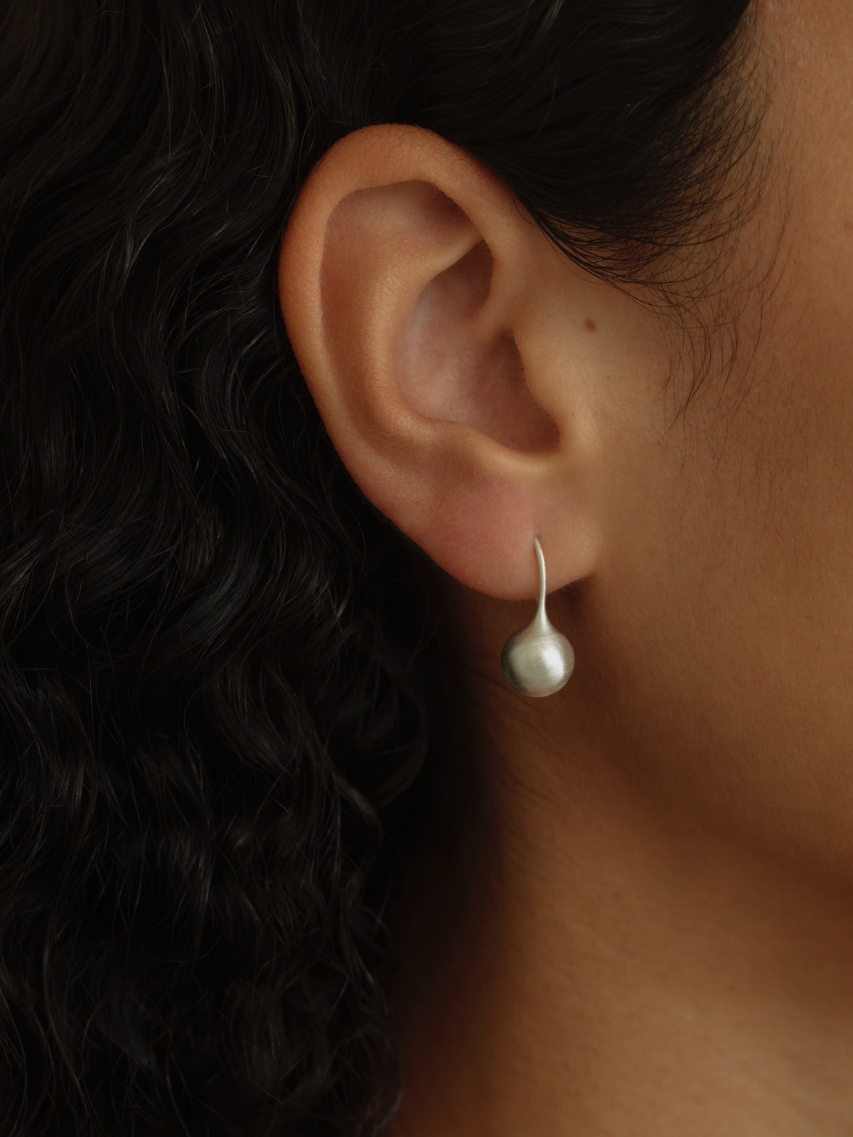 A close-up of a person's ear wearing a single silver drop earring with a teardrop-shaped white pearl, set against dark curly hair.