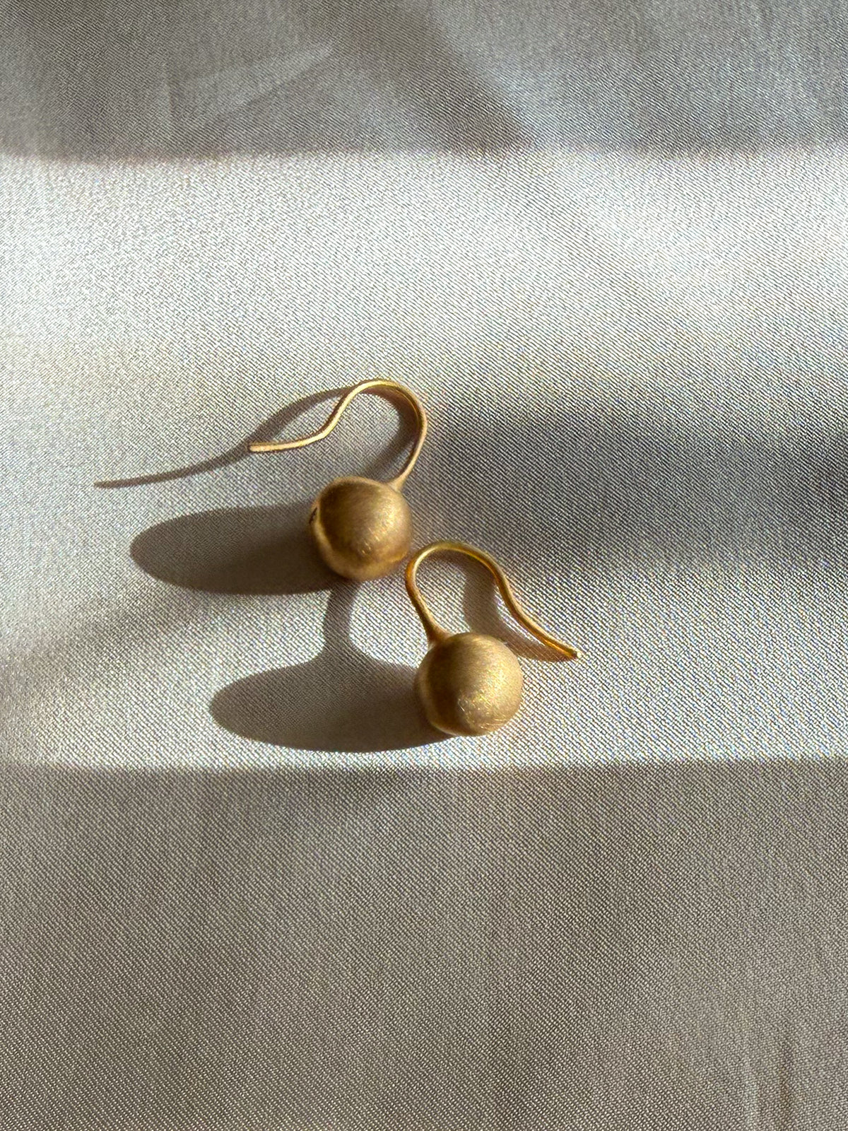 A pair of gold drop earrings with textured spherical beads and curved hook backs, resting on a textured beige fabric under warm, directional sunlight.