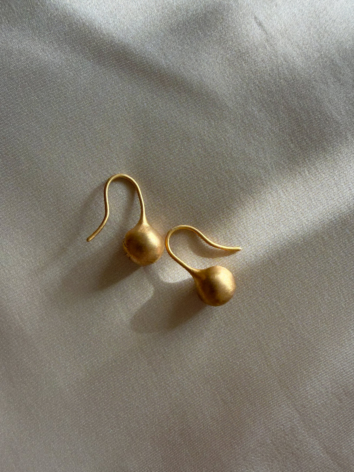 A pair of gold dangle earrings with teardrop-shaped drops and curved hooks, resting on a textured off-white fabric background with soft shadows.