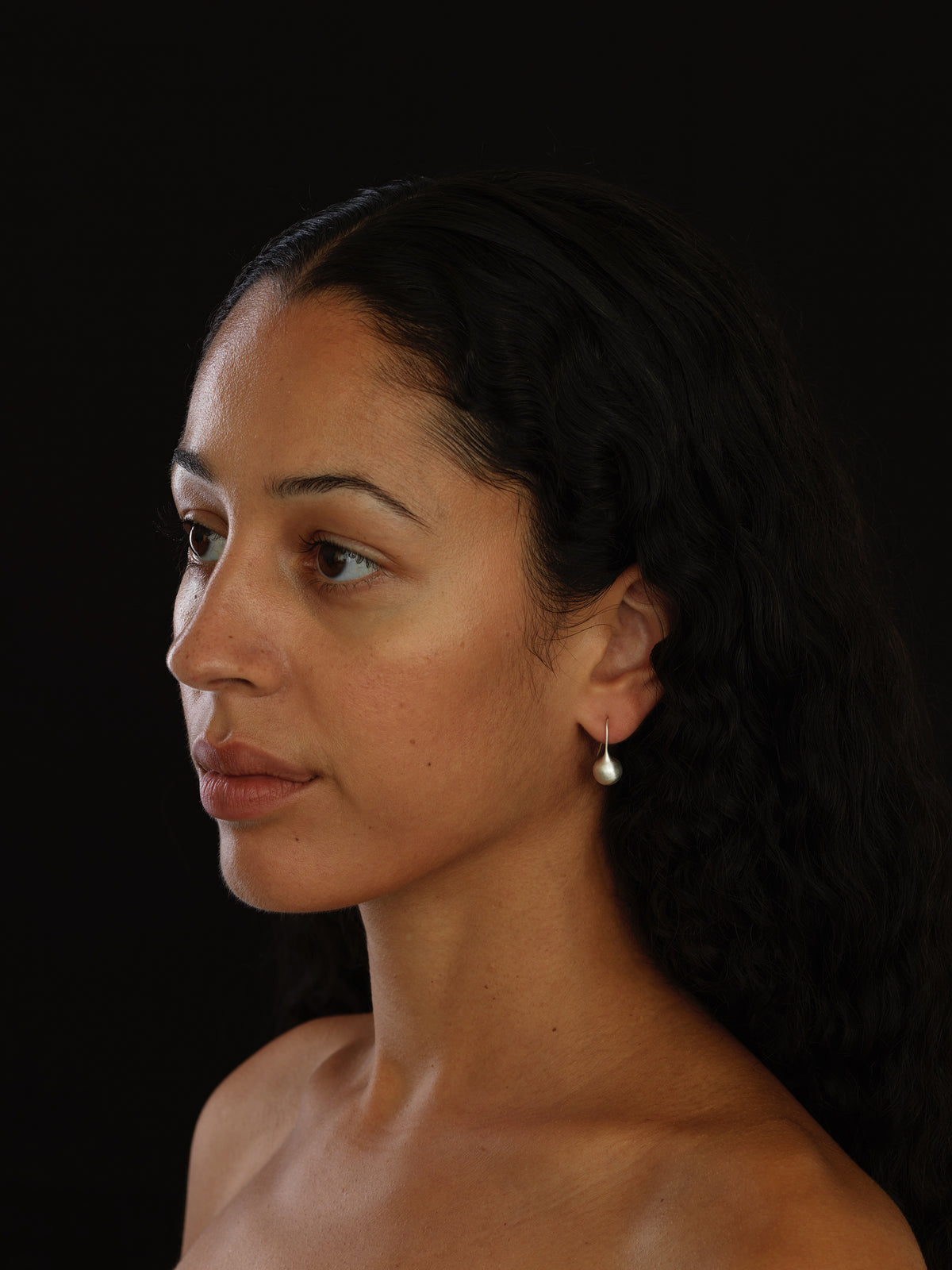 A close-up side profile of a woman with dark hair wearing a small, dangling pearl-like earring against a solid black background.